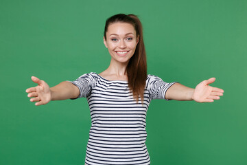 Smiling joyful attractive young brunette woman 20s wearing striped casual clothes posing standing reach out stretch hands looking camera isolated on green color wall background, studio portrait.