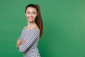 Side view of smiling attractive young brunette woman 20s wearing striped casual clothes posing standing holding hands crossed looking aside isolated on green color wall background, studio portrait.