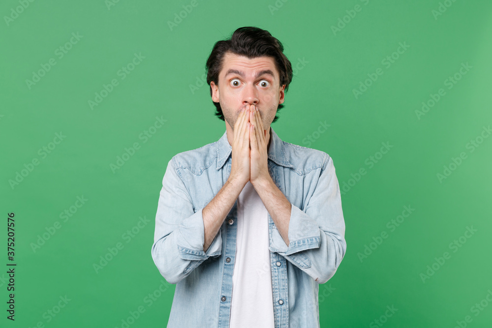 Wall mural Shocked amazed young brunet man 20s wearing casual clothes white t-shirt denim shirt posing standing covering mouth with hands looking camera isolated on green color wall background studio portrait.