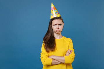 Displeased dissatisfied offended young brunette woman 20s in yellow casual clothes birthday hat posing celebrating holding hands crossed looking camera isolated on blue background studio portrait.