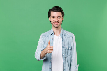 Smiling funny young brunet man 20s wearing casual clothes white t-shirt denim shirt posing standing showing thumb up looking camera isolated on green color wall background studio portrait.