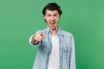 Angry irritated young brunet man 20s wearing casual clothes white t-shirt denim shirt posing standing pointing index finger on camera screaming swearing isolated on green background studio portrait.