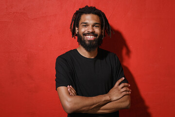 Smiling young african american man guy with dreadlocks 20s wearing black casual t-shirt posing holding hands crossed folded looking camera isolated on bright red color wall background studio portrait.