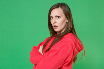 Obraz premium Side view of displeased perplexed young brunette woman 20s wearing bright red casual streetwear hoodie posing holding hands crossed looking camera isolated on green color background studio portrait.