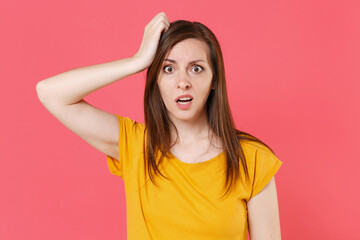 Shocked perplexed bewildered worried puzzled young brunette woman 20s wearing yellow casual t-shirt posing put hand on head looking camera isolated on pink color wall background studio portrait.