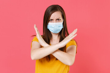 Young woman in yellow t-shirt sterile face mask to safe from coronavirus virus covid-19 2019-ncov during quarantine showing stop gesture with crossed hands isolated on pink background studio portrait.