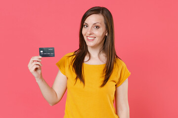 Smiling beautiful attractive young brunette woman 20s in yellow casual t-shirt posing standing holding in hands credit bank card looking camera isolated on pink color wall background studio portrait.