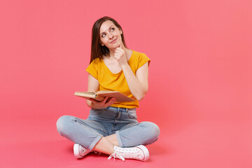 Full length portrait of pensive young brunette woman 20s in yellow casual t-shirt posing sitting on floor reading book put hand prop up on chin looking up isolated on pink color background studio.
