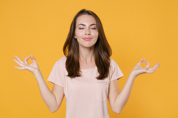 Smiling young brunette woman 20s wearing pastel pink casual t-shirt posing hold hands in yoga gesture relaxing meditating trying to calm down isolated on yellow color wall background studio portrait.