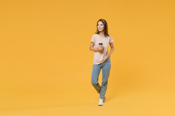 Full length portrait of smiling young brunette woman 20s wearing pastel pink casual t-shirt posing hold paper cup of coffee or tea looking aside isolated on bright yellow color wall background studio.