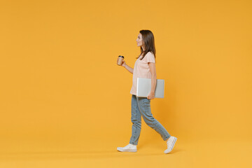Full length side view portrait of smiling young brunette woman 20s in pastel pink casual t-shirt posing hold laptop pc computer paper cup of coffee or tea isolated on yellow color background studio.