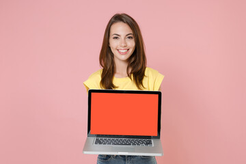 Naklejka premium Smiling young brunette woman 20s wearing yellow casual t-shirt hold laptop pc computer with blank empty screen mock up copy space looking camera isolated on pastel pink background studio portrait.