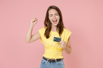 Happy joyful young brunette woman in yellow casual t-shirt posing standing holding credit bank card doing winner gesture keeping eyes closed isolated on pastel pink wall background studio portrait.