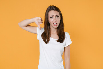 Displeased worried dissatisfied young brunette woman 20s wearing white blank empty design casual t-shirt posing showing thumb down looking camera isolated on yellow wall background studio portrait.