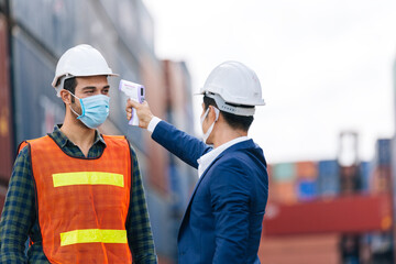 Manager or engineer foreman using infrared thermometer measure temperature to construction site staff worker at shipping container warehouse. men at work wearing face mask prevent Covid-19 infection
