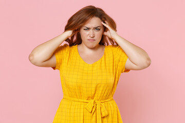 Displeased tired puzzled young redhead plus size body positive female woman girl 20s in yellow dress posing put hands on head having headache isolated on pastel pink color background studio portrait.