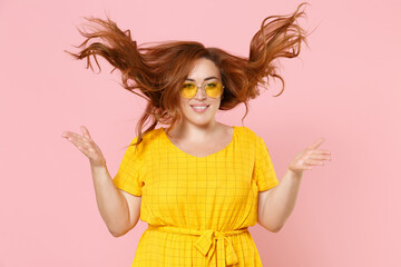 Smiling pretty young redhead plus size body positive female woman 20s in yellow dress eyeglasses posing having fun throwing up fluttering hair isolated on pastel pink color background studio portrait.