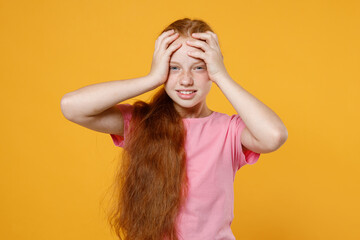 Displeased worried little ginger redhead kid girl 12-13 years old in pink casual t-shirt posing put hands on head looking camera isolated on bright yellow color background children studio portrait.