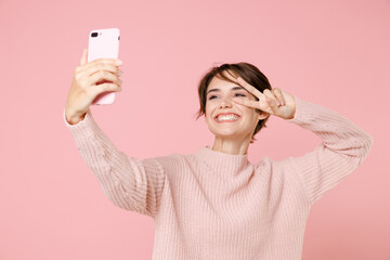 Smiling young brunette woman girl in knitted casual sweater posing isolated on pastel pink background studio portrait. People lifestyle concept. Doing selfie shot on mobile phone showing victory sign.