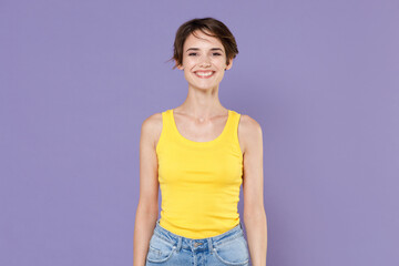 Smiling beautiful young brunette woman girl wearing yellow casual tank top posing isolated on pastel violet wall background studio portrait. People sincere emotions lifestyle concept. Looking camera.