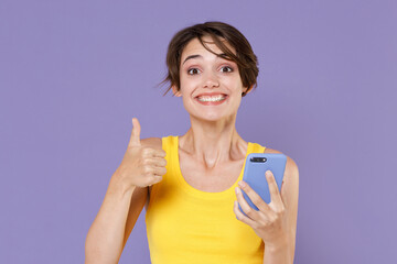 Smiling young brunette woman girl in yellow casual tank top posing isolated on pastel violet background studio portrait. People emotions lifestyle concept. Using mobile cell phone, showing thumb up.