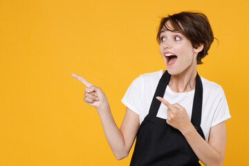 Surprised young female woman 20s barista bartender barman employee in white casual t-shirt apron pointing index fingers aside on mock up copy space isolated on yellow color background studio portrait.