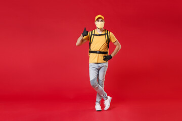 Full length delivery employee man in yellow cap face mask gloves t-shirt uniform thermal food bag backpack work courier service during quarantine coronavirus covid-19 virus isolated on red background.