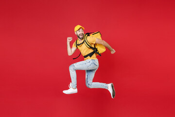 Full length delivery employee man guy male 20s in yellow cap t-shirt uniform thermal food bag backpack work courier service during quarantine covid-19 virus jumping isolated on red background studio.