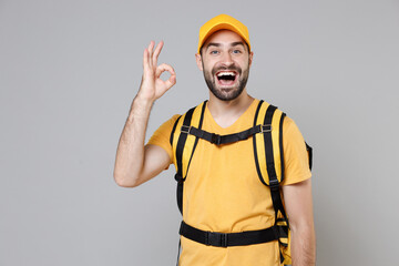 Delivery employee man guy male 20s in yellow cap t-shirt uniform thermal food bag backpack work courier service during quarantine coronavirus covid-19 virus, posing isolated on gray background studio.