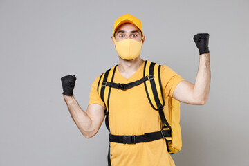 Delivery employee man guy in yellow cap t-shirt uniform face mask gloves thermal food bag backpack work courier service during quarantine coronavirus covid-19 virus isolated on gray background studio.