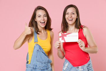 Two cheerful funny young brunette women friends 20s wearing casual denim clothes posing hold in hand gift certificate showing thumb up isolated on pastel pink colour background, studio portrait.