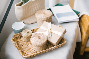 A gift box tied with a beige satin ribbon with a bow, straw baskets with lids on a wicker tray, and...