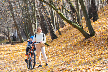 Obraz premium Happy mother leads a bicycle with a child strapped in the back in the autumn park.