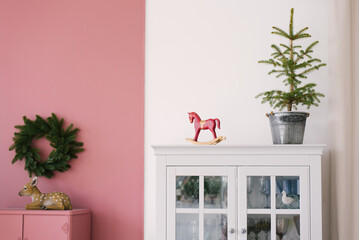Christmas tree in a metal bucket, vintage wooden red toy horse on a white dresser in the interior of the living room, decorated for Christmas and New Year. A fawn and a wreath over a pink Cabinet