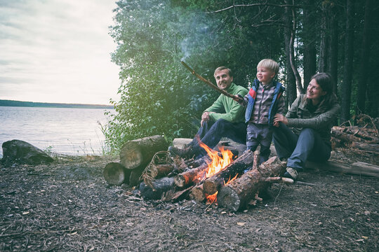 Family Weekend Outdoor. Father, Mother And Child Sitting Near Bonfire In Forest Near Lake. Local Travel, Camping, Hiking Lifestyle.