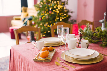 White plates, champagne glasses, tangerines in the serving of a festive dinner for Christmas, Christmas eve against the background of Christmas tree lights