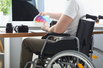 Man in white T-shirt and green pant sit in wheelchair and hold multi-colored palette in his hand. There is monitor, camera, len and keyboard on table.
