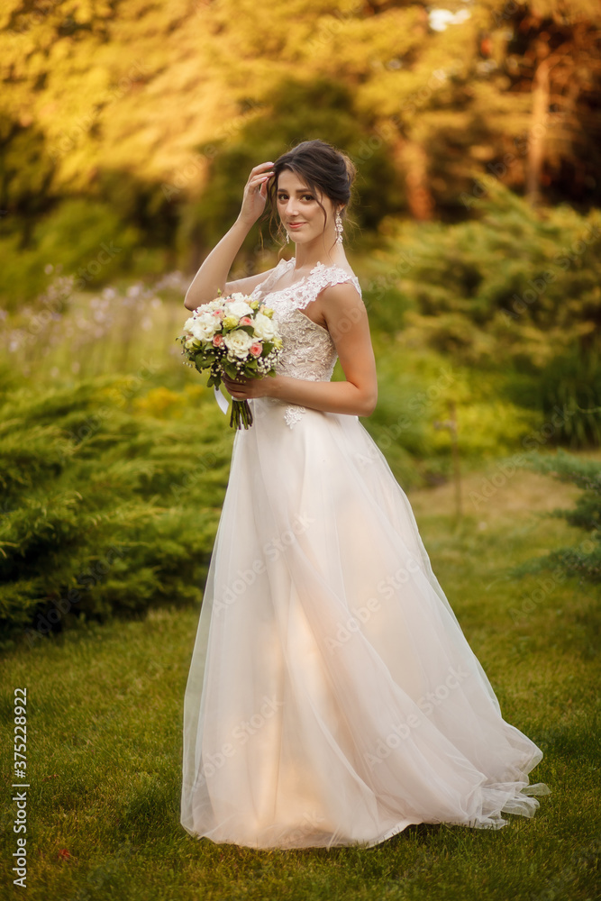 Wall mural A beautiful bride in a white dress straightening her hair holds a wedding bouquet in her hand. Bride on a walk on a warm autumn day. Portrait of a happy wedding bride on their wedding day