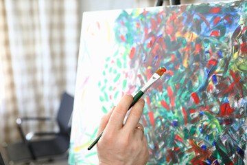 Artist hold brush in his hand. Close up male hand draw picture with multi-colored paint. Painting with multi-colored brush stroke stand on easel.