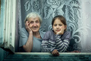 An old woman with a small granddaughter together look out of the window of the house.