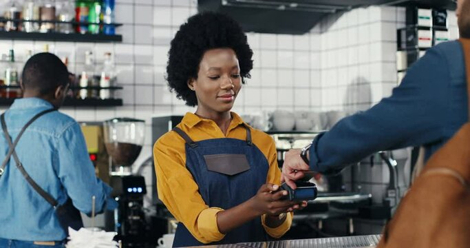 Multiethnic bartender woman and client man. Caucasian male paying with paypass, credit card. Mixed-races. Passing payment at counter. Pay for drink or food in cafe. African American waitress.