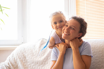 Young mother with little kid daughter hugging, having fun, in good loving mood smiling looking at camera, sitting on light sofa at home. Mother's Day love family, parenthood childhood concept