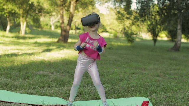 Athletic child girl in VR headset helmet making fitness workout exercises with dumbbells in park