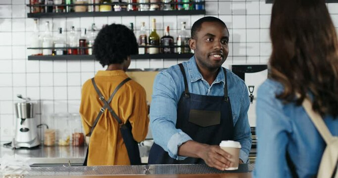 African American waiter serving coffee and talking with Caucasian woman at counter in cafe. Man barista having nice talk with customer. Male bartender chatting with female client. Mixed-races.