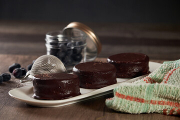 Chocolate alfajor on a white platter, with fresh blueberries, sugar and several. on a wooden table and dark background