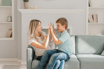 Naklejka na ściany i meble Mom and kid are laughing together in cozy home. They are holding each other with joy while relaxing on sofa. Parent is playfully touching boy feet and he is enjoying it