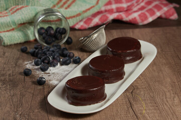 Chocolate alfajor on a white platter, with fresh blueberries, sugar and several. on a wooden table and dark background