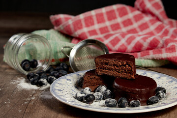 Chocolate alfajor on a white platter, with fresh blueberries, sugar and several. on a wooden table and dark background