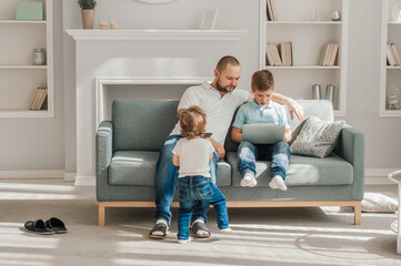 Happy young father sit on couch using laptop relax with preschooler son holding smartphone have fun together, smiling dad and little boy child enjoy weekend at home rest on sofa busy with gadgets