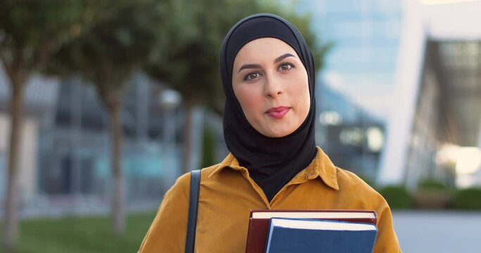 Portrait shot of young Arabic smiled beautiful woman in black hijab standing at street with textbooks. Good-looking muslim female student in traditional Arab headscarf at city holding books. Outside.